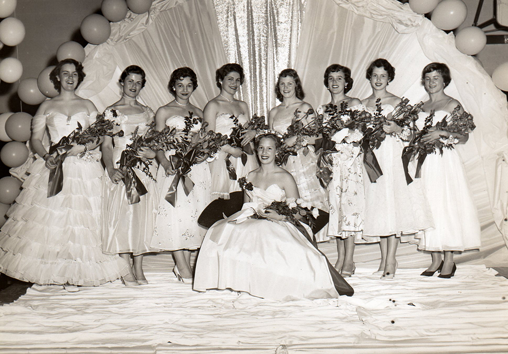 Portrait of prom queen seated in center and eight girls standing behind her in front of festive backdrop